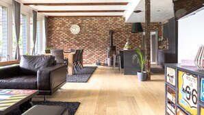 A modern living room with a brick wall, a fireplace, a dining area, and a black leather sofa. - Holiday Home 'Beautiful View' in the Harz with Mountain View, Private Terrace and Wi-Fi (Walkenried)