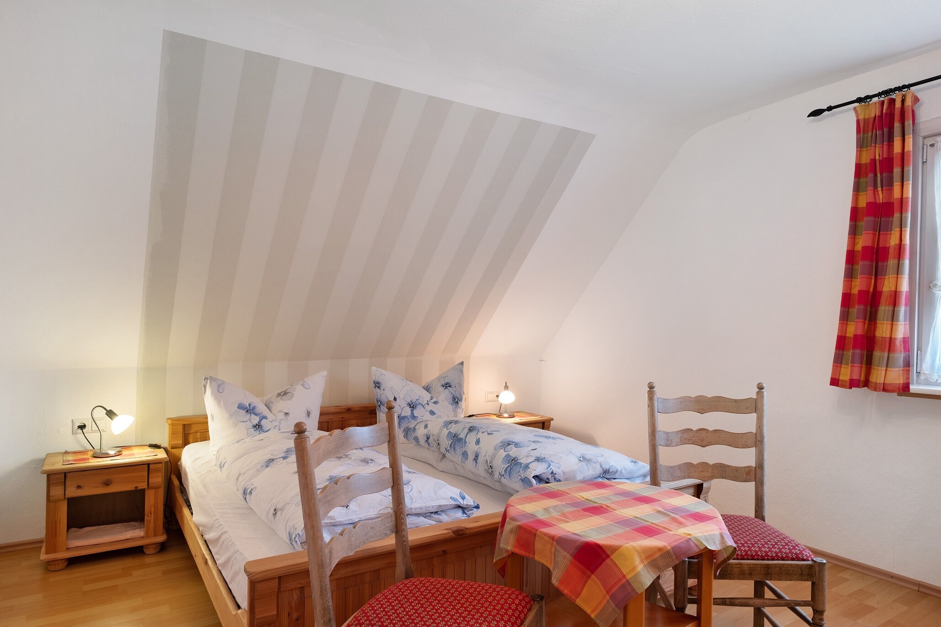 A bedroom with a sloped ceiling, a bed with floral bedding, a wooden chair, a small table with a checkered tablecloth, and a nightstand with a lamp.
