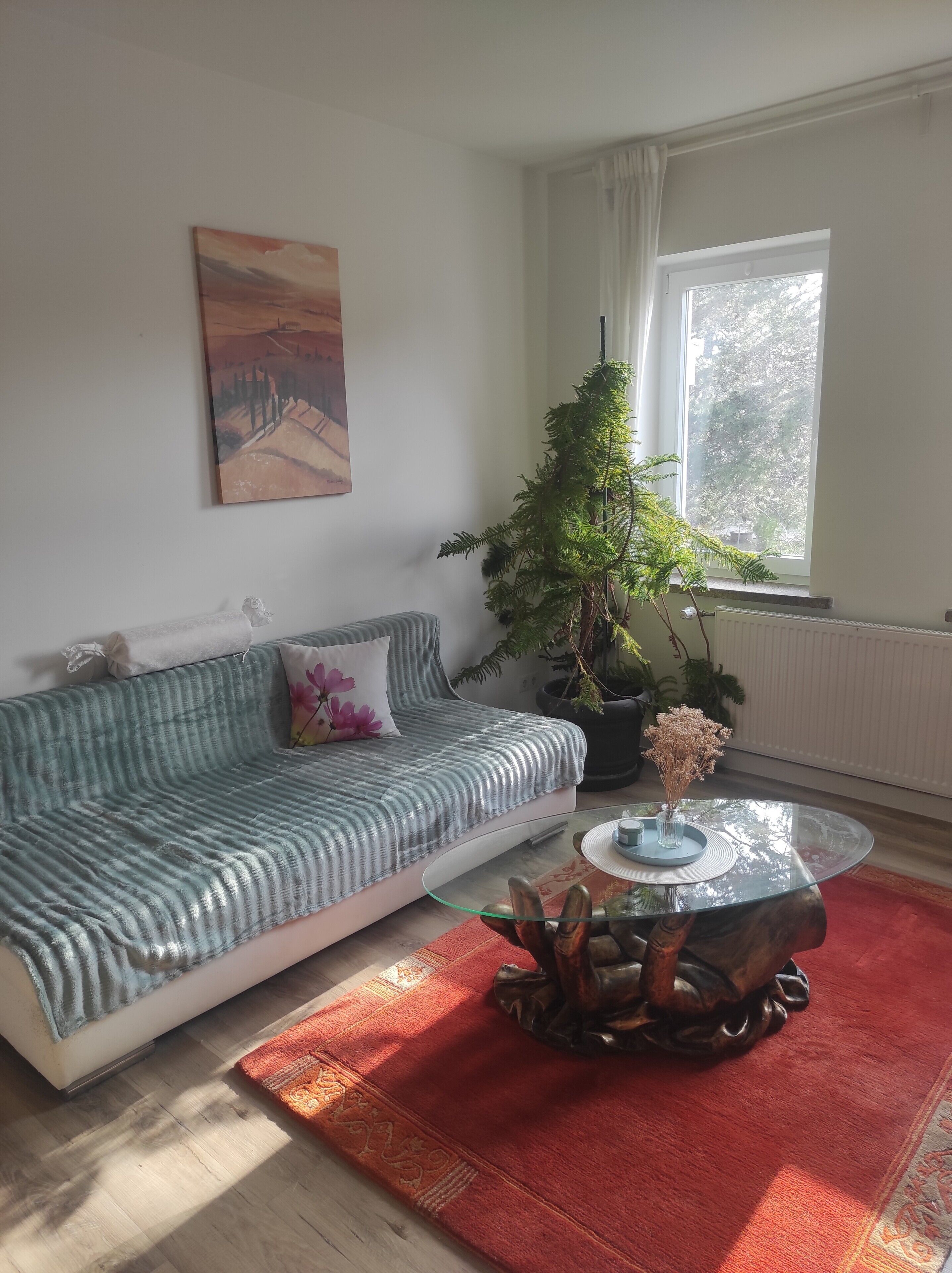 Una sala de estar con un sofá, una mesa de centro de vidrio, una planta en maceta y un cuadro en la pared.