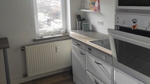 Una cocina con armarios blancos, horno empotrado, microondas y ventana con vista a árboles.