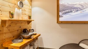 Un intérieur en bois avec une étagère en bois où sont posées des bouteilles et des verres, un lavabo en pierre et une horloge.