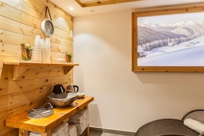 A wooden interior with a wooden shelf holding bottles and glasses, a stone basin, and a clock.