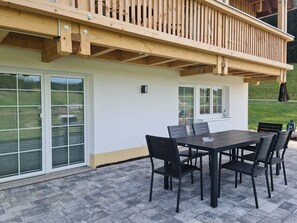 Een overdekt terras met een tafel en stoelen, een houten terras en een witte gevel.