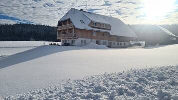 Ein großes, mehrstöckiges Holzhaus mit geneigtem Dach, umgeben von verschneitem Gelände und einem bewaldeten Hintergrund.