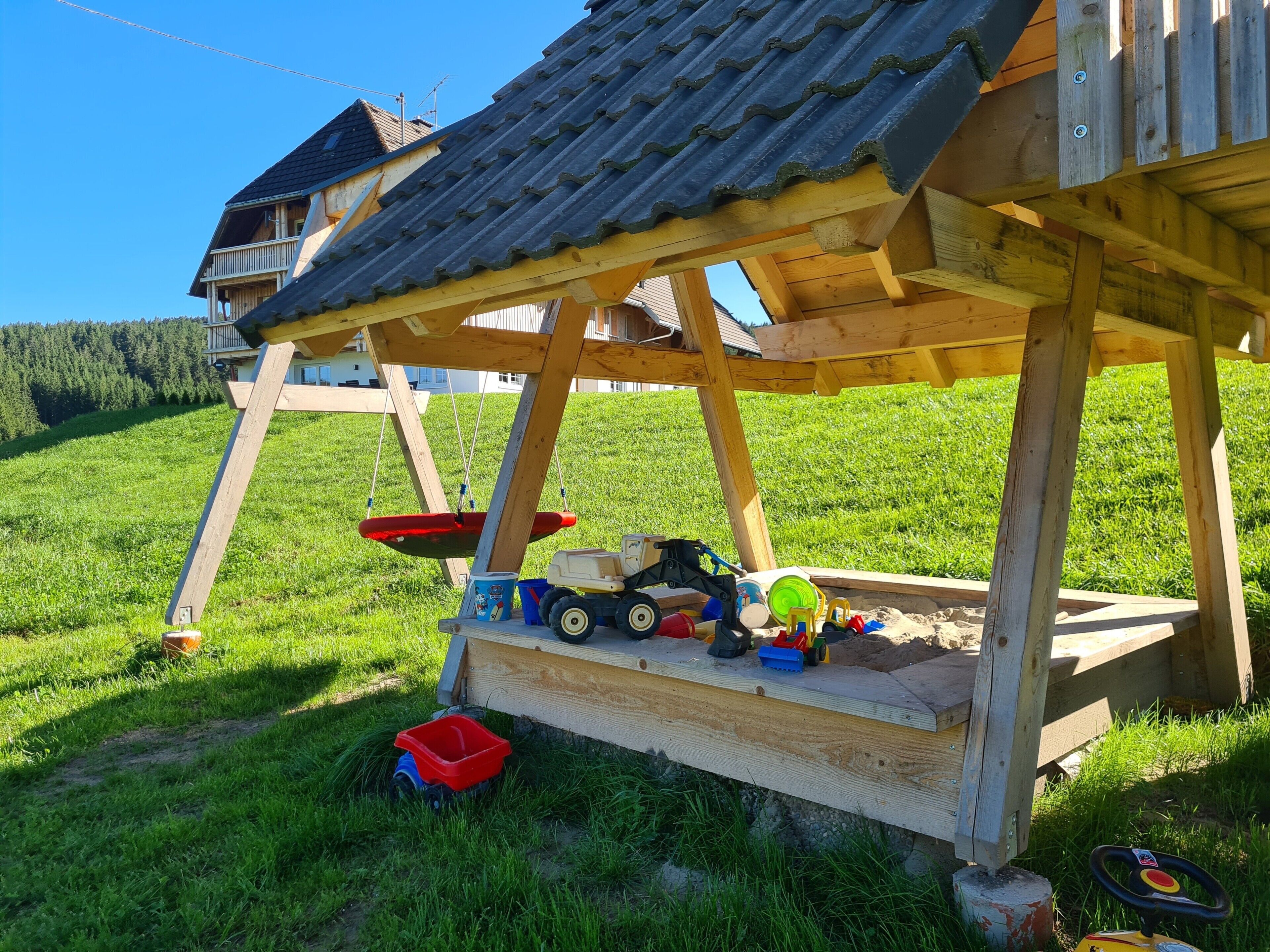 Ein hölzernes Spielhäuschen mit geneigtem Dach, einer Schaukel und verschiedenen Spielsachen auf dem Boden.