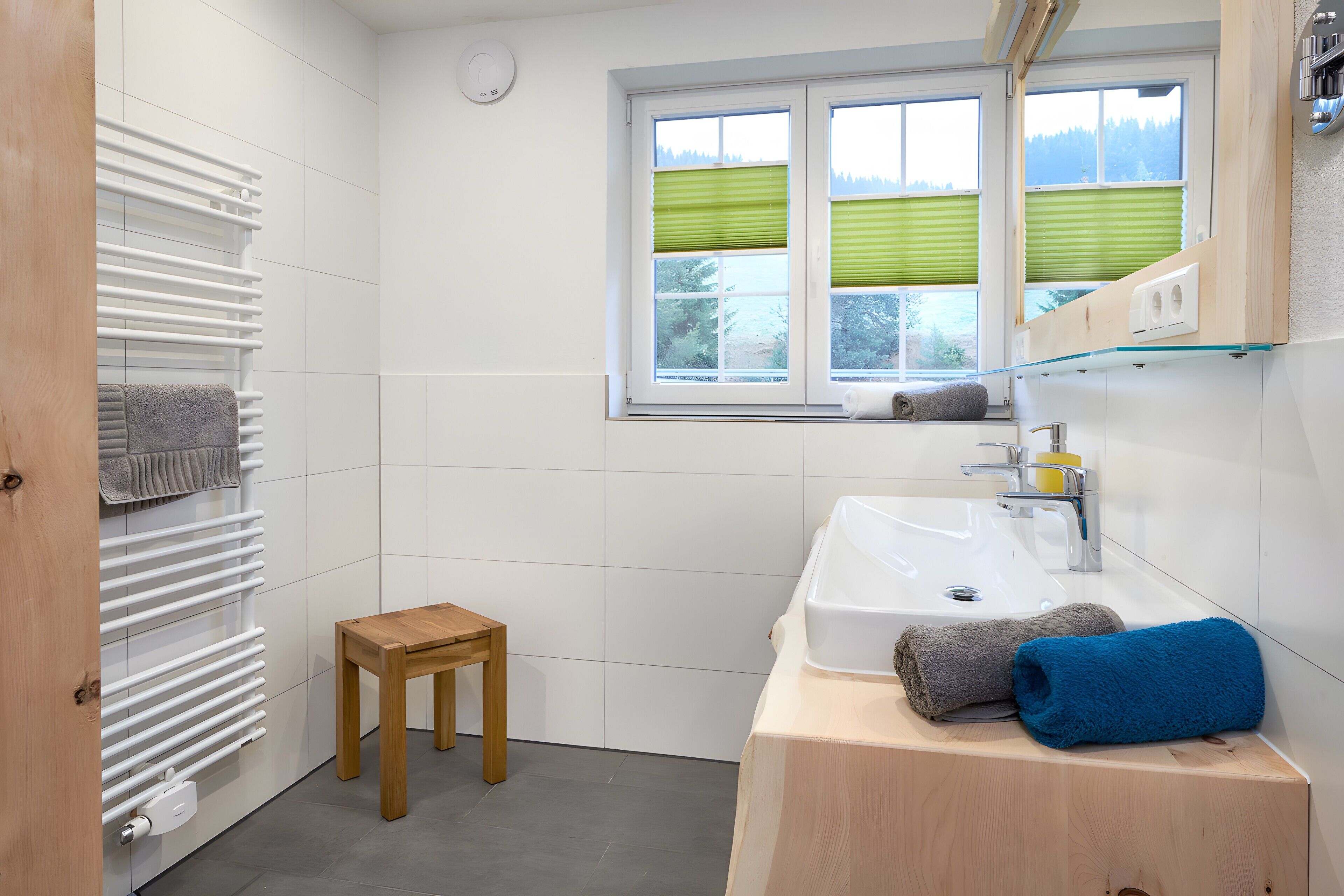 A bathroom with a white radiator, a wooden stool, a sink with a mirror, and a window with green blinds.
