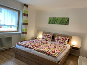 Ein Bett mit gemusterten Textilien, eine Nachttischlampe, eine Uhr und ein Fenster mit Vorhängen.