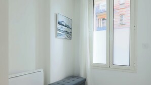 A corner room with a radiator, a grey bench, a window with a view of buildings, and a framed picture on the wall. - Flat 1BDR/2P Roland Garros/Parc des (Boulogne-Billancourt)