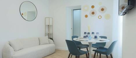 A modern living room with a white sofa, a round mirror, and a dining area with a white table and blue chairs.