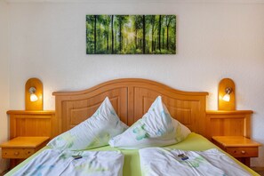 Un dormitorio con cabecera de madera, dos mesitas de noche con lámparas, una colcha verde y un cuadro con motivos forestales sobre la cama.