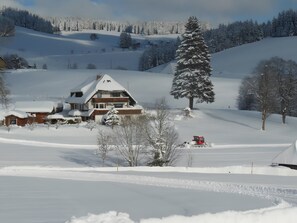 A snow-covered landscape with a house, a large evergreen tree, and a red vehicle.