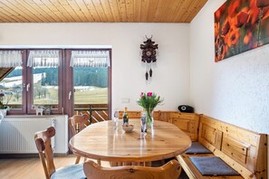 A wooden dining table with chairs, a bench, a cuckoo clock, and a painting of flowers. - Holiday Apartment 'Schöne Aussicht, 60sqm' with Shared Garden, Balcony and Wi-Fi (Titisee-Neustadt)