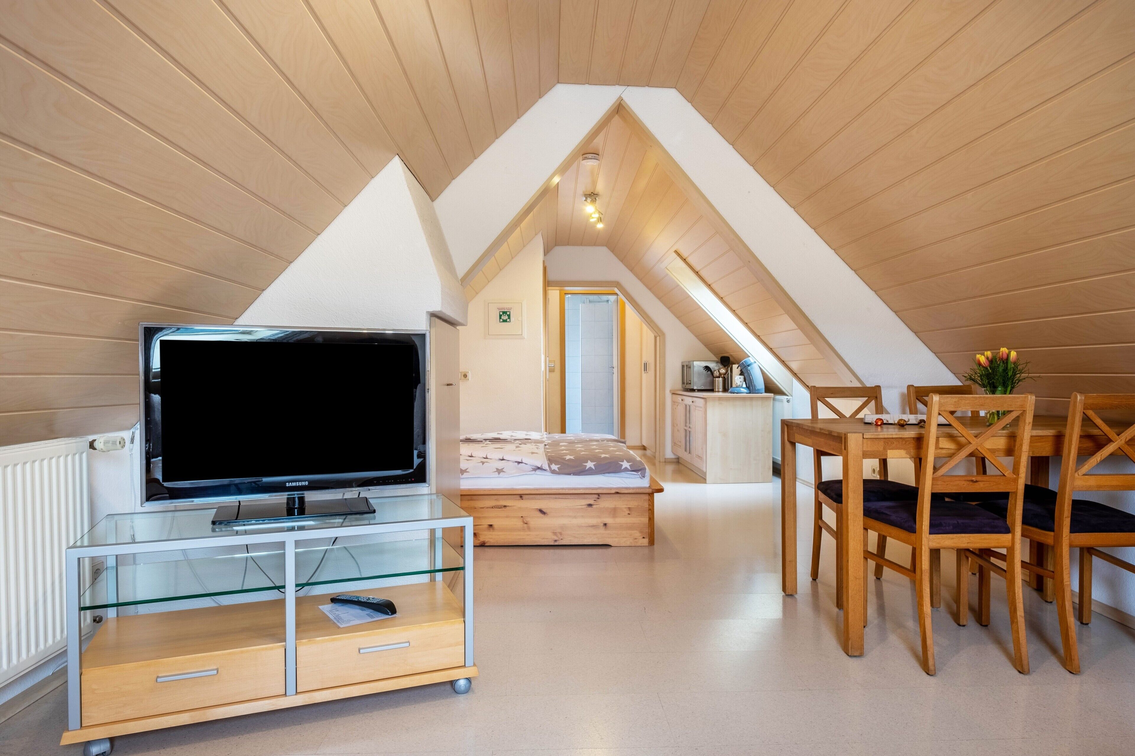 Ein Loft-Zimmer mit Fernseher auf einer Glastruhe, einem Bett mit Bettdecke mit Sternenmuster, einem Essbereich mit Holzsesseln und einer Küchenzeile mit weißer Arbeitsplatte.