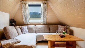 Une pièce chaleureuse avec un plafond en bois, une grande fenêtre avec des rideaux, un canapé avec des coussins, une petite table avec une bouteille et des verres, et un ventilateur fixé au mur.