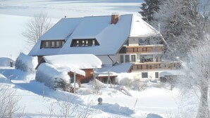 Une maison à deux étages, dotée d’un toit en pente recouvert de neige, et entourée d’un paysage enneigé.