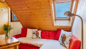 Une pièce chaleureuse avec un plafond en bois, un canapé rouge, un coussin blanc, une lampe et une fenêtre donnant sur un beau paysage.