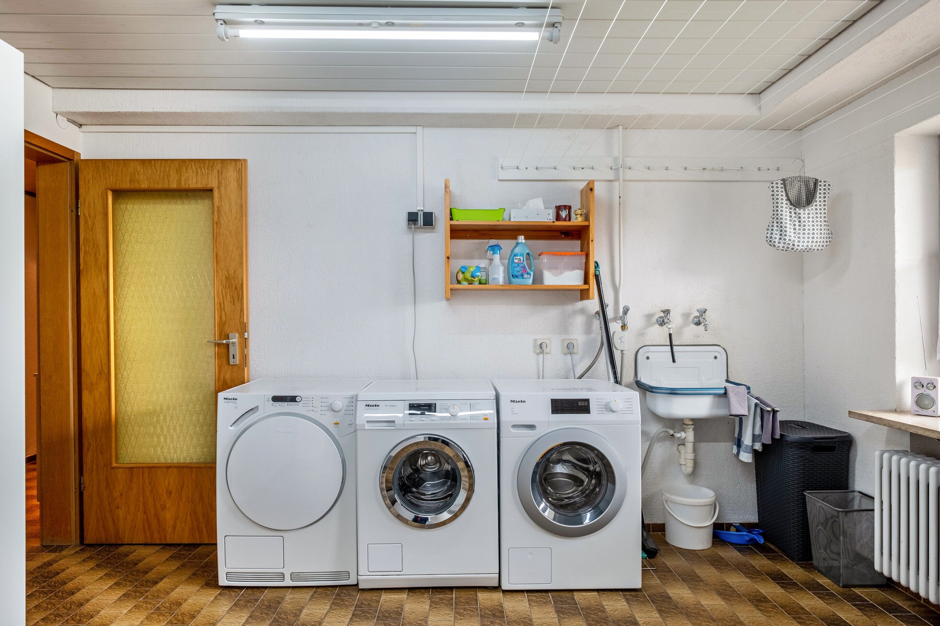 A laundry room with three front-loading washing machines, a sink, a radiator, and a wooden door.