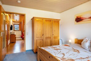 Une chambre avec une armoire en bois, un lit recouvert d’une couvre-lit à motifs floraux, une table de chevet et un tableau accroché au mur.