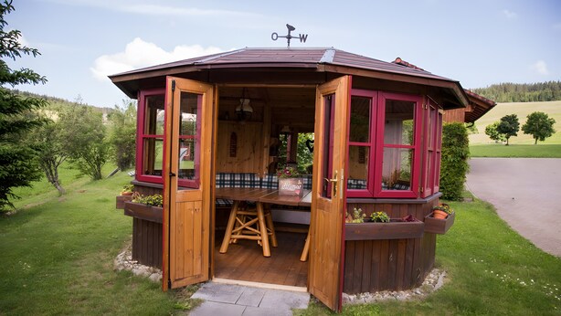 Un belvédère en bois, avec une table et des chaises, entouré de verdure.