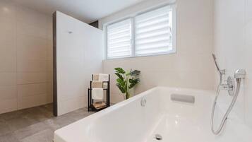 A modern bathroom with a bathtub, a shower, and a window with blinds.