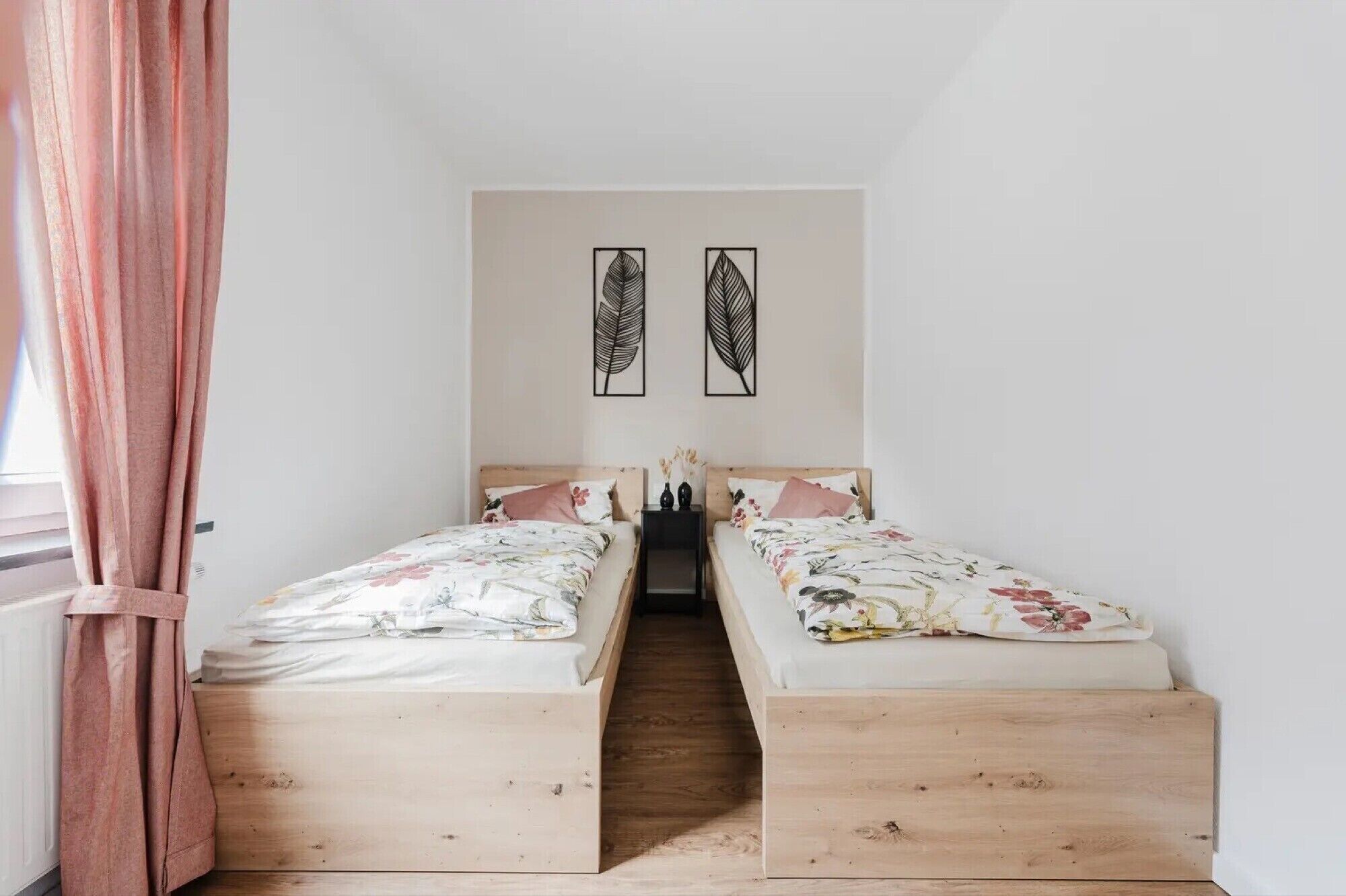 A bedroom with two single beds, a wooden floor, a window with pink curtains, and two leaf-patterned wall art pieces.