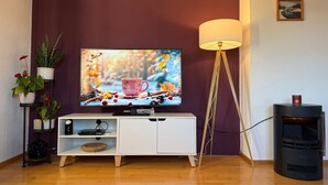 Una sala de estar con televisor sobre un mueble blanco, una lámpara de pie, chimenea y un cuadro colgado en la pared.