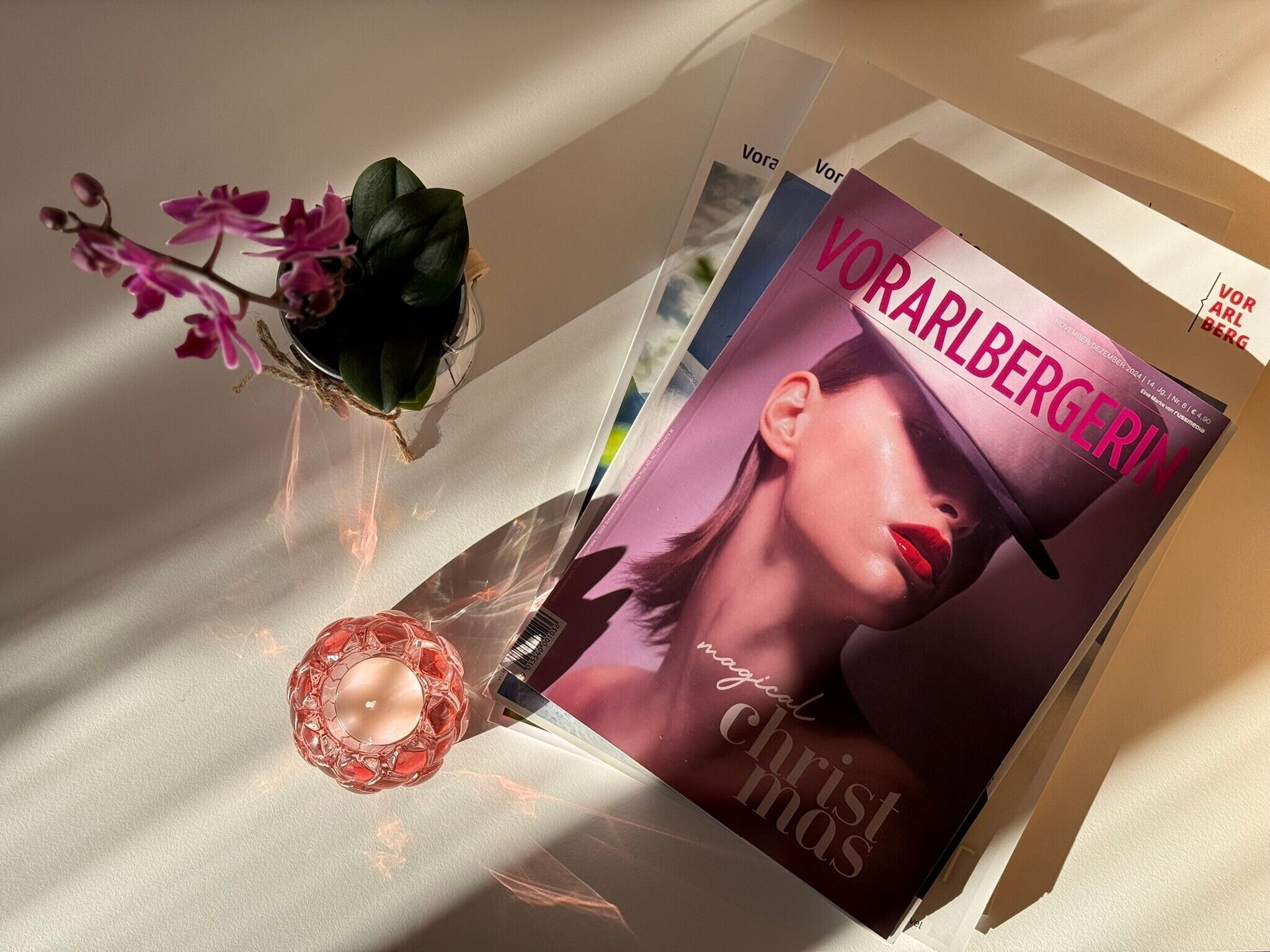 Una revista con un retrato de mujer, una vela de cristal rosa y un florero con flores moradas.