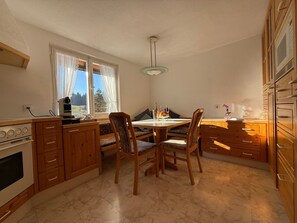 A kitchen with wooden cabinets, a dining table with chairs, a built-in oven, and a window with curtains.