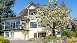 Ein zweistöckiges Gebäude mit Balkon, einer Treppe zum Eingang und einem gepflegten Garten mit verschiedenen Blumen.