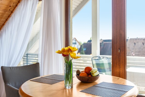 A dining table with a vase of yellow flowers, a bowl of fruit, and a view of buildings through large windows.