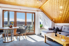 A modern living room with a sofa, a dining table with chairs, and a balcony with a view.