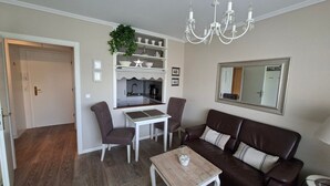 Una sala de estar con un sofá de cuero marrón, una mesa de centro de madera, una mesa de comedor con sillas, una cocina con pileta y alacenas, y un candelabro.