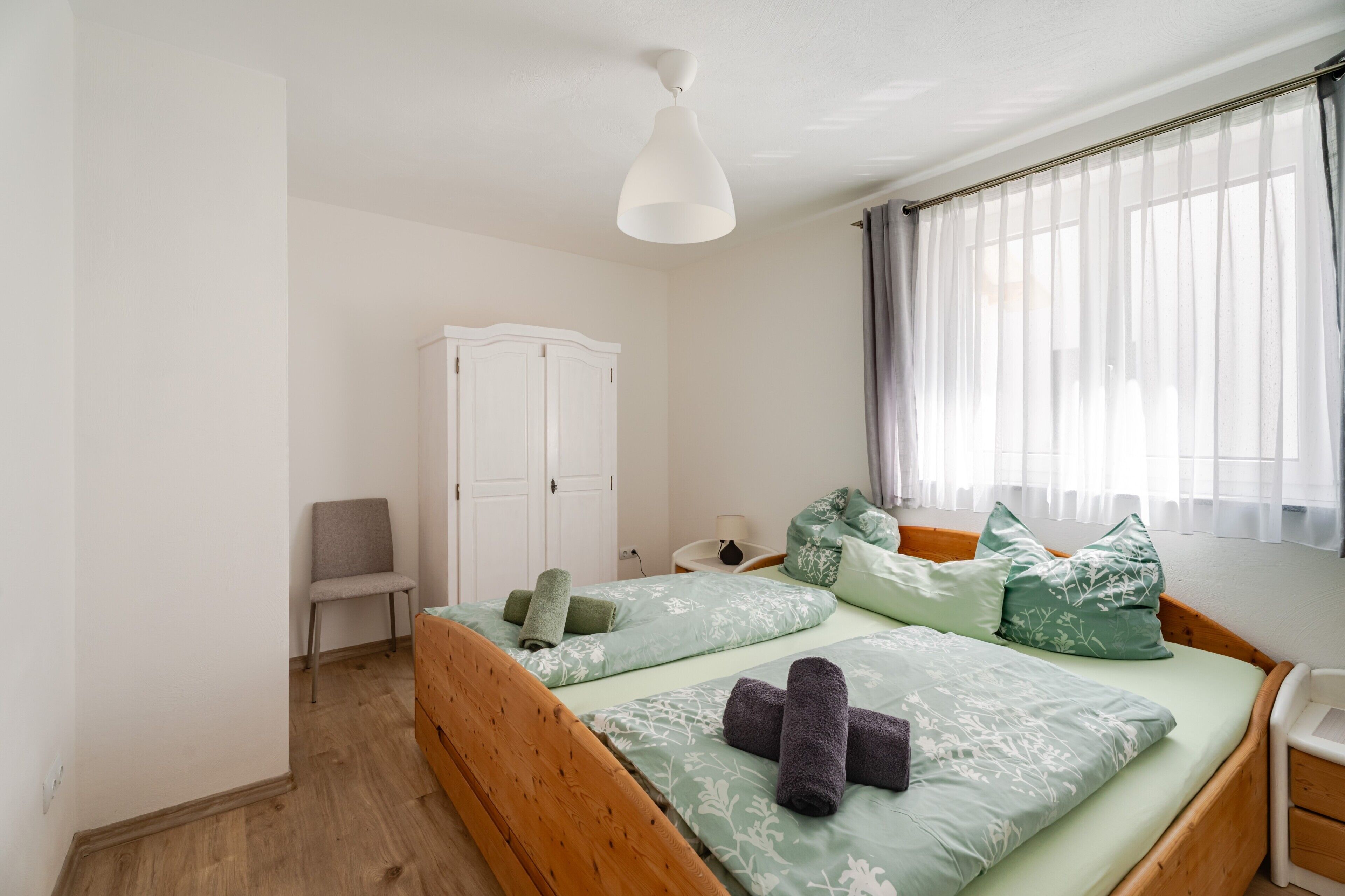 Une chambre à coucher avec un lit en bois, une armoire blanche, une chaise et une fenêtre avec des rideaux.