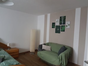 Ein Zimmer mit einem grünen Sofa, einer Stehlampe, einer Wand mit gerahmten Pflanzenabbildungen und einem Holzboden.