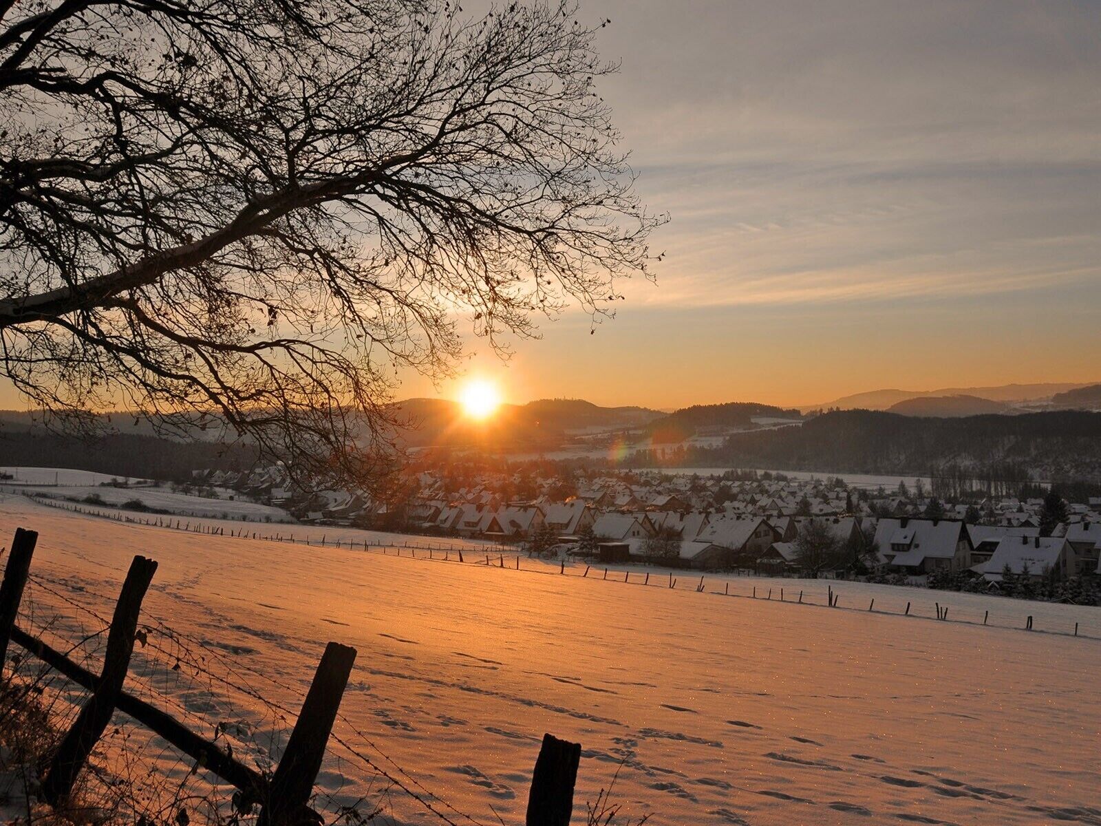 A snowy landscape with a sunset, a fence, and houses in the distance.