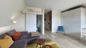 Un salon moderne comprenant un canapé, une petite table basse, un fauteuil bleu et un coin cuisine avec des placards.