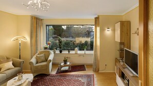 A living room with a sofa, armchair, TV, and a large window with a view of greenery. - Holiday Apartment 'Auszeit' with Private Terrace, Shared Garden and Wi-Fi (Arnsberg)