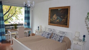 Een slaapkamer met een bed, een bureau, een stoel, een kroonluchter, een schilderij en een raam met gordijnen.