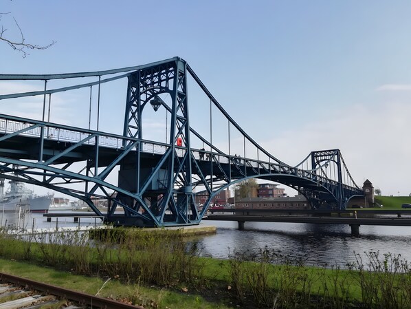 A large blue bridge with a pedestrian walkway, a river below, and a ship docked in the background.