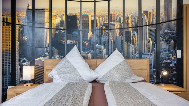 Una camera d'albergo con un letto, cuscini, un comodino, una lampada e vista sulla città attraverso ampie finestre.