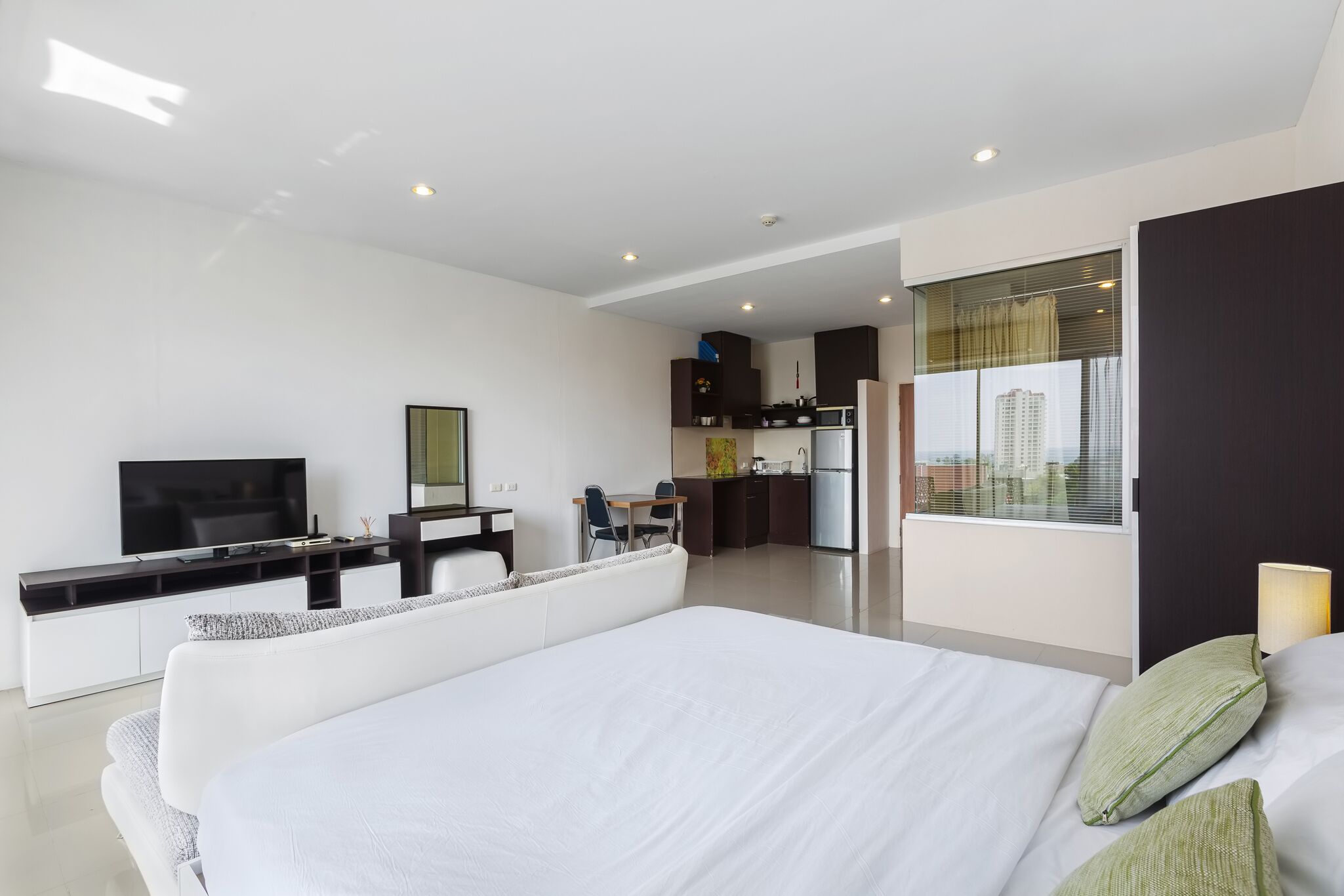 A modern hotel room with a large bed, a TV, a desk, and a kitchenette in the background.