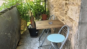 Una pequeña zona exterior con mesa y silla de metal, una planta en maceta y suelo de hormigón.