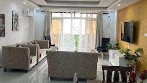 Ein modernes Wohnzimmer mit einer Sofagarnitur, einem Esstisch mit Unterlagen und einem Deckenventilator, sowie einem an der Wand befestigten Fernseher.