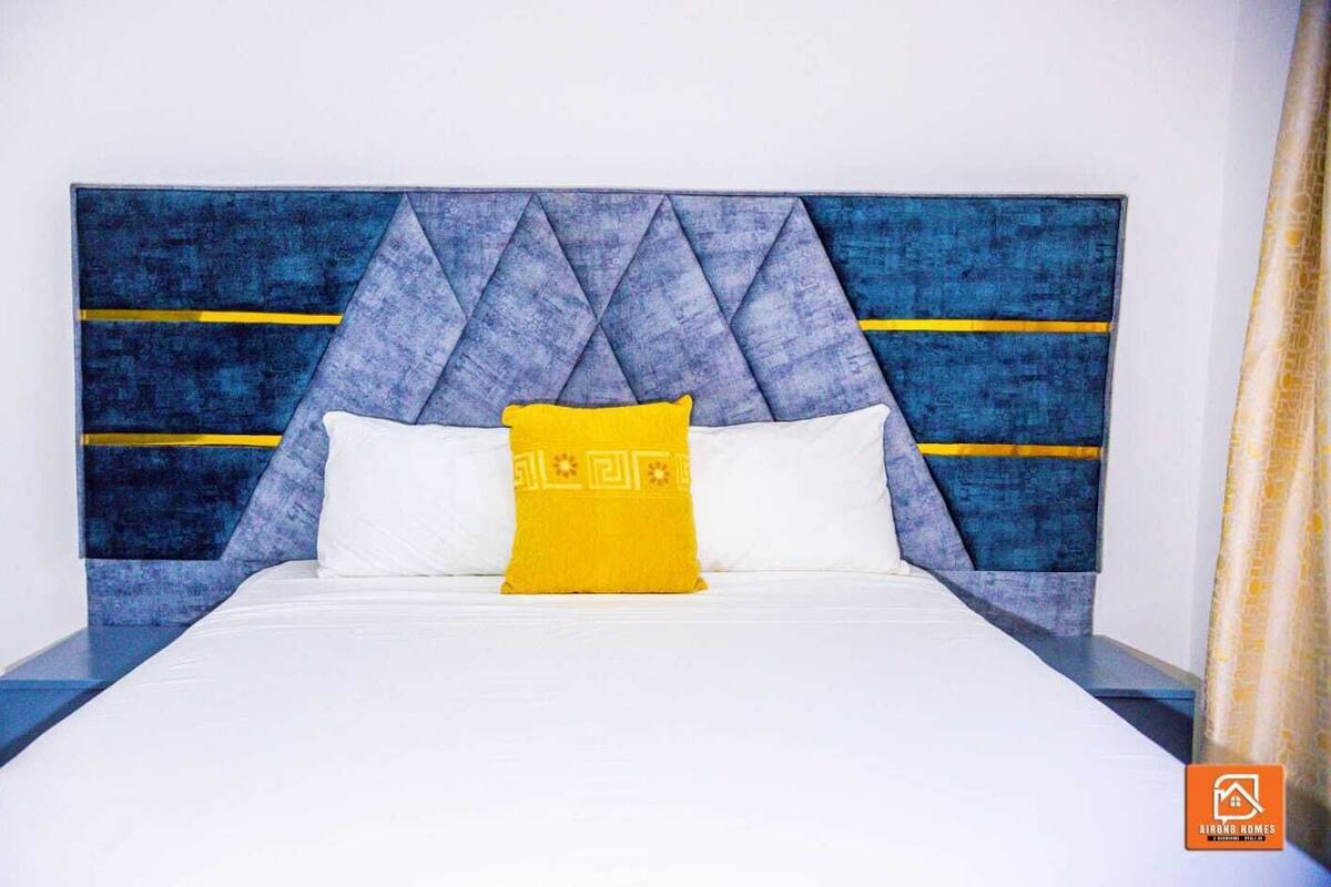 Una cama con cabecera azul, ropa de cama blanca y almohada amarilla.