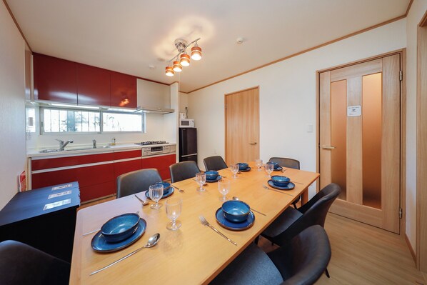 Un comedor con una mesa de madera dispuesta para cuatro personas, con platos azules, vasos y cubiertos. Se aprecia una cocina roja y blanca al fondo.
