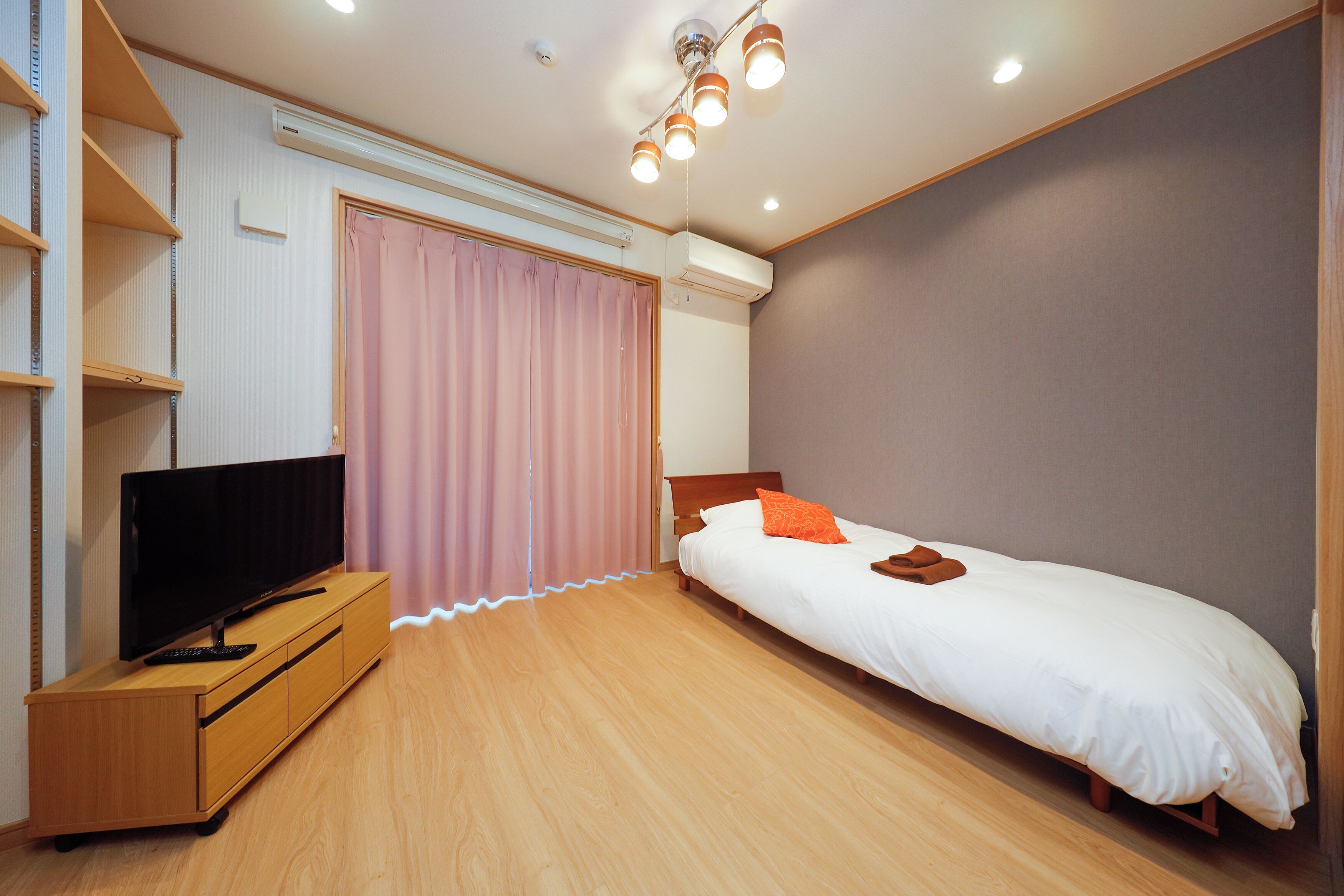 Une chambre à coucher avec un lit, un téléviseur posé sur un meuble en bois, une fenêtre avec des rideaux roses et un luminaire au plafond.