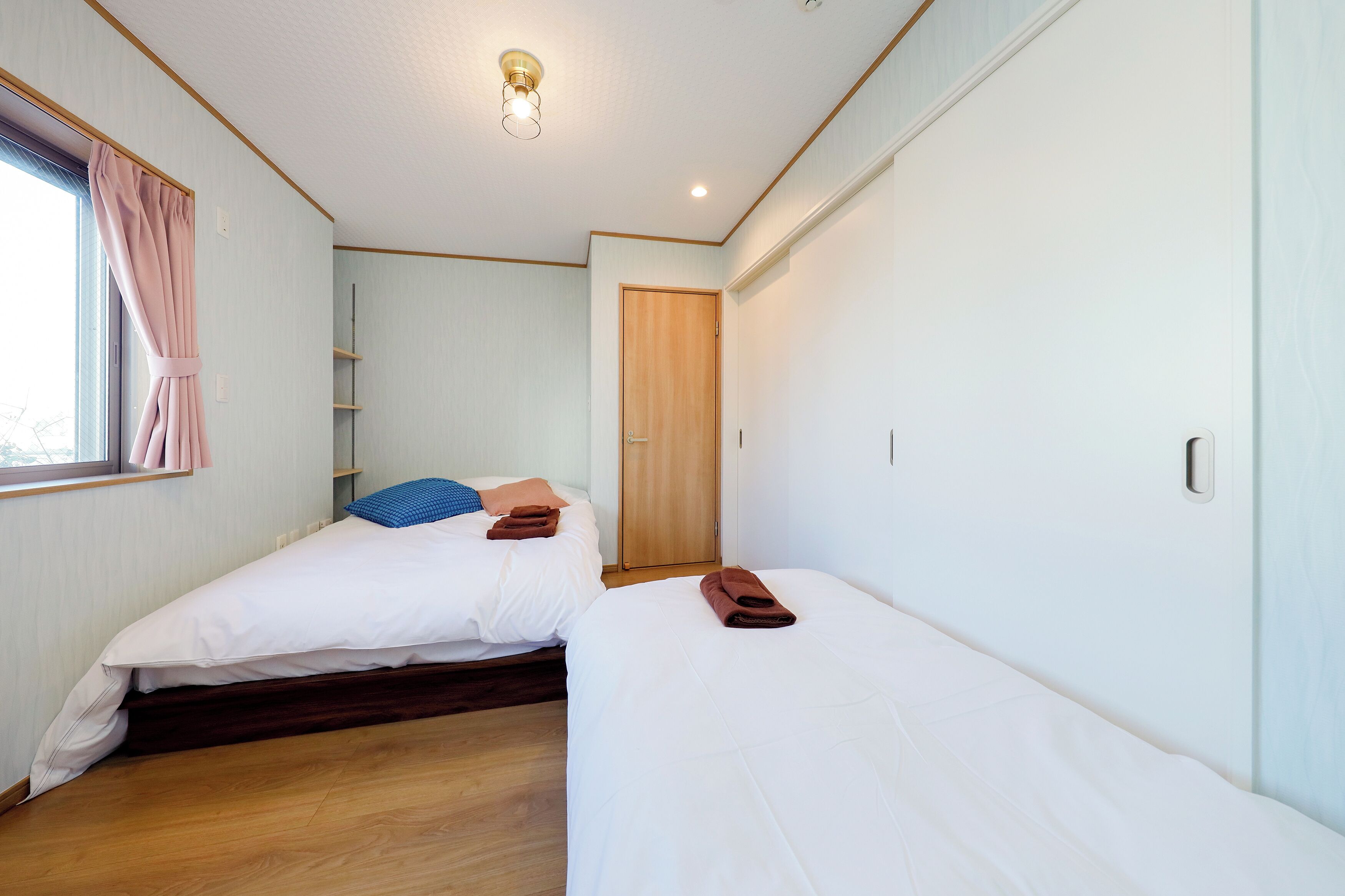 A hotel room with two single beds, a wooden door, a window with curtains, and a ceiling light.