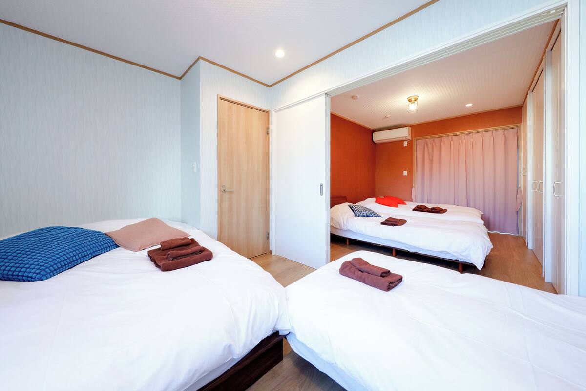 A hotel room with two beds, each with white bedding and brown towels. The room has a wooden floor, white walls, and a wooden door.