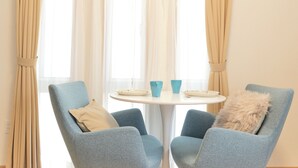 A room with two blue armchairs, a round white table with two blue cups and plates, and beige curtains.
