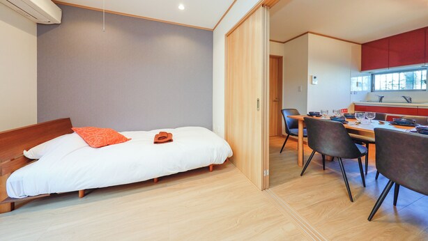 A modern room with a bed, a dining table with chairs, and a kitchen area visible through an open door.